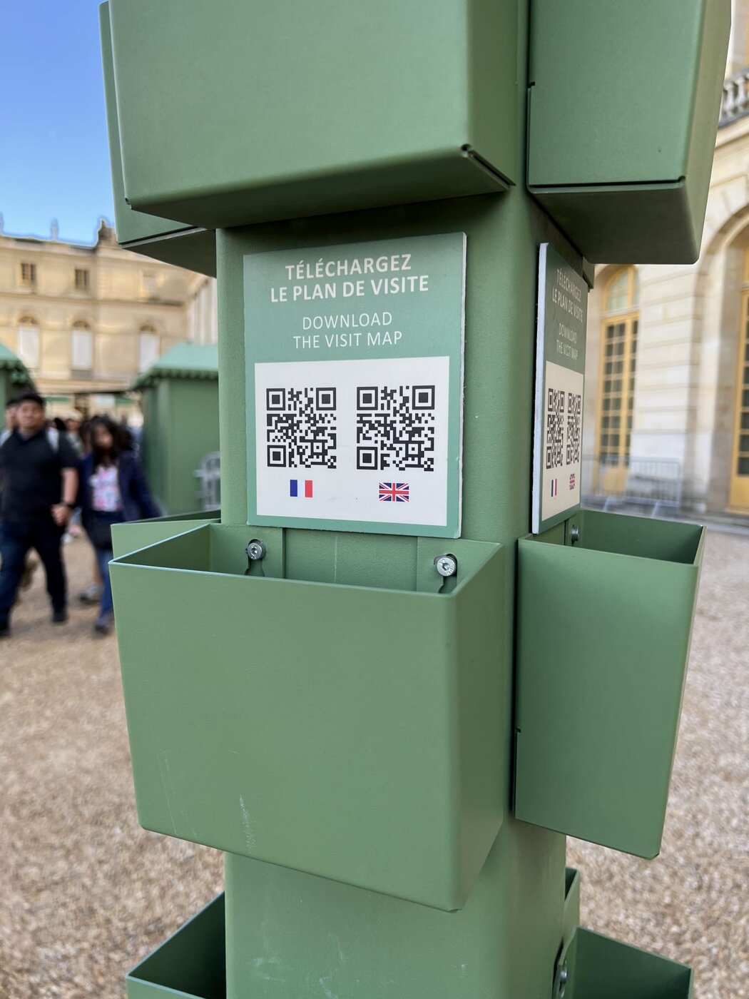 A QR code kiosk at the Château de Versailles with French and English flag-labeled codes for downloading the visit map PDF